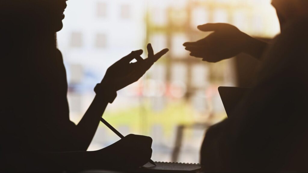 Silhouette of two people talking with one person holding a pencil about to write in a notebook with the modern office as a background.