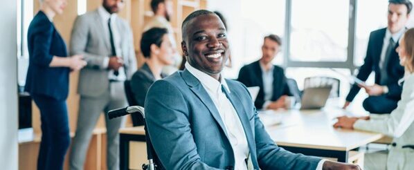 A young businessman in a wheelchair is smiling while in a business meeting in the board room with his colleagues in the background.